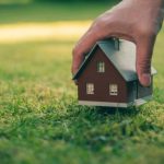 Concept of selling a house. A hand is holding a model house above green meadow.
